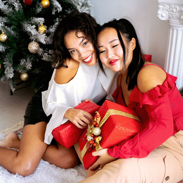 Two women exchange gifts and smile at the camera in front of a Christmas tree. For Giving the Gift of Great Smiles blog post.