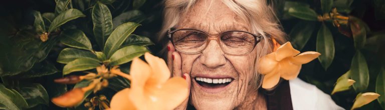 Elderly woman smiling brightly with a full set of beautiful teeth among summer flowers. Banner for Skip Buyer's Remorse blog post.