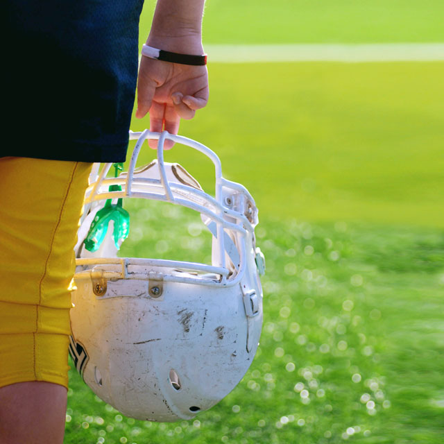 Football player holds their helmet with attached athletic mouthguard while looking across a field.