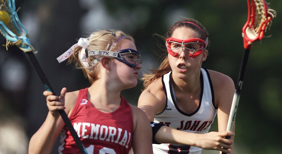 Two lacrosse players in full uniform, including athletic mouthguards, run across the field while fighting for control of the ball