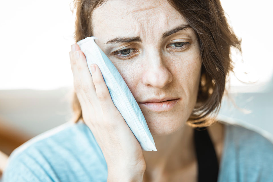 A woman with mouth pains holds an ice pack to her cheek. Looking to replace missing teeth.