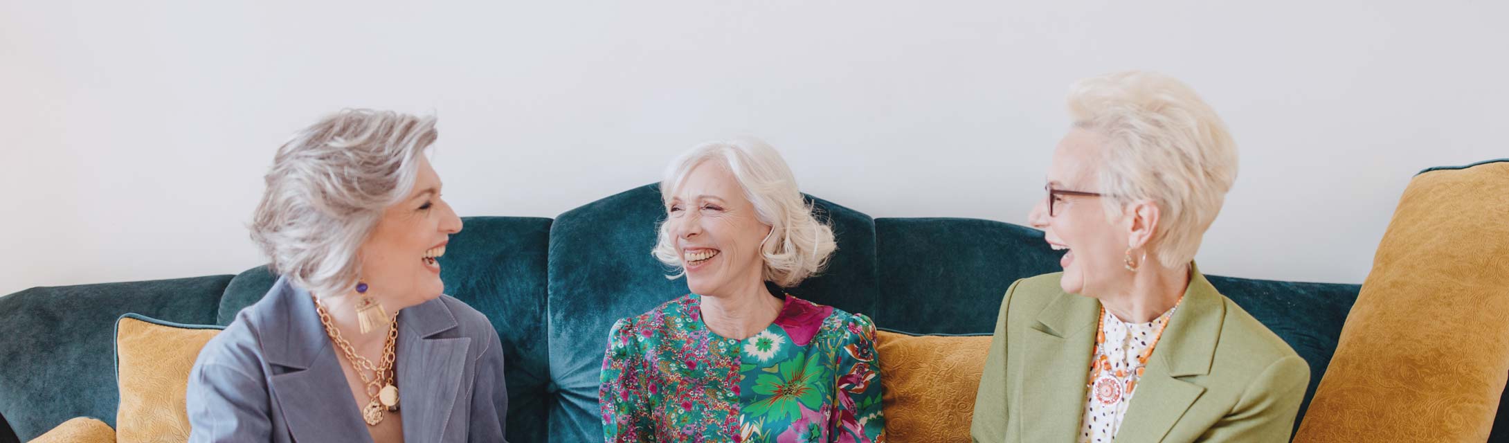 Three older women laugh while sitting on a couch. Banner image for helping your dentures last blog post.