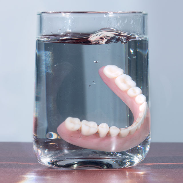 Traditional dentures being dropped into a glass of water. Helping your dentures last through proper care is essential.