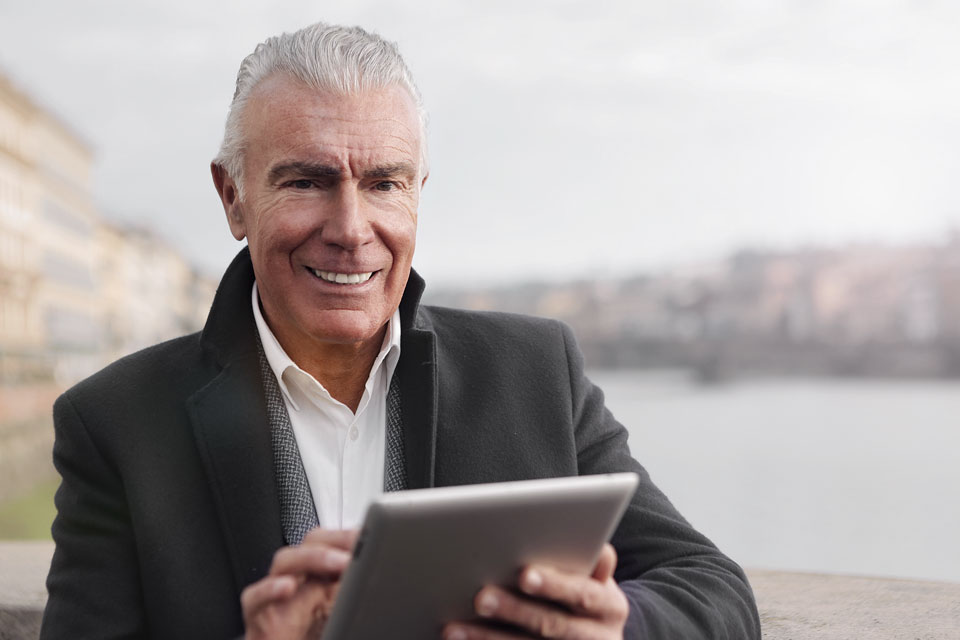 An older gentleman smiles while using a tablet. Helping your dentures last means sharing more smiling memories with loved ones.