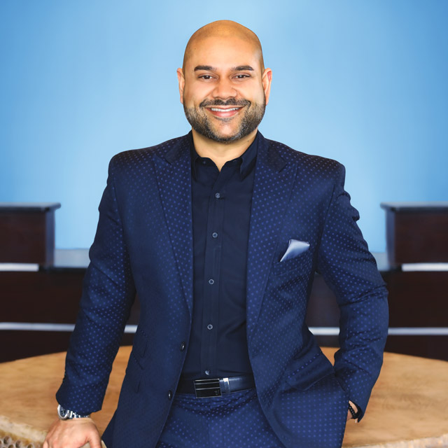 Dr. Neal Patel smiles wearing a blue suit in the front lobby of his dental practice Infinite Smiles. Image for Destination Dentistry blog post.